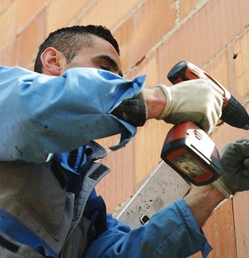 a man in a blue jacket and gloves uses a power drill on a brick wall